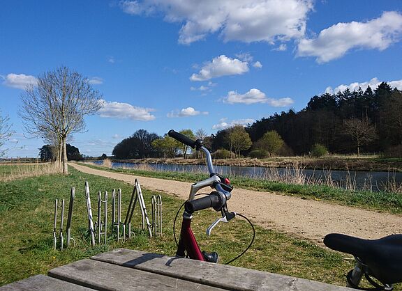 Ein Blick auf den Elbe-Lübeck-Kanal. Im Hintergrund ist ein Fahrradlenker, angelehnt an einen Tisch zu sehen. Blauer Himmel mit Schäfchenwolken